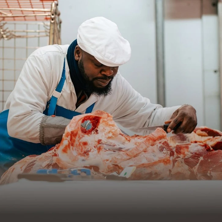 Portrait professionnel montrant deux hommes d'âges différents (un homme d'âge mûr et un homme plus jeune) côte à côte dans un environnement de boucherie artisanale ou atelier de vente de viande. Ils portent des tenues professionnelles appropriées (chemise, tablier de boucher ou tenue commerciale). Le cadre inclut des éléments évoquant leur métier (vitrines réfrigérées, planches de découpe, ou produits de qualité en arrière-plan). L'atmosphère doit être chaleureuse et professionnelle, mettant en avant la transmission familiale et la complémentarité des générations. Photo en éclairage naturel ou professionnel, composition équilibrée, sans filigrane.